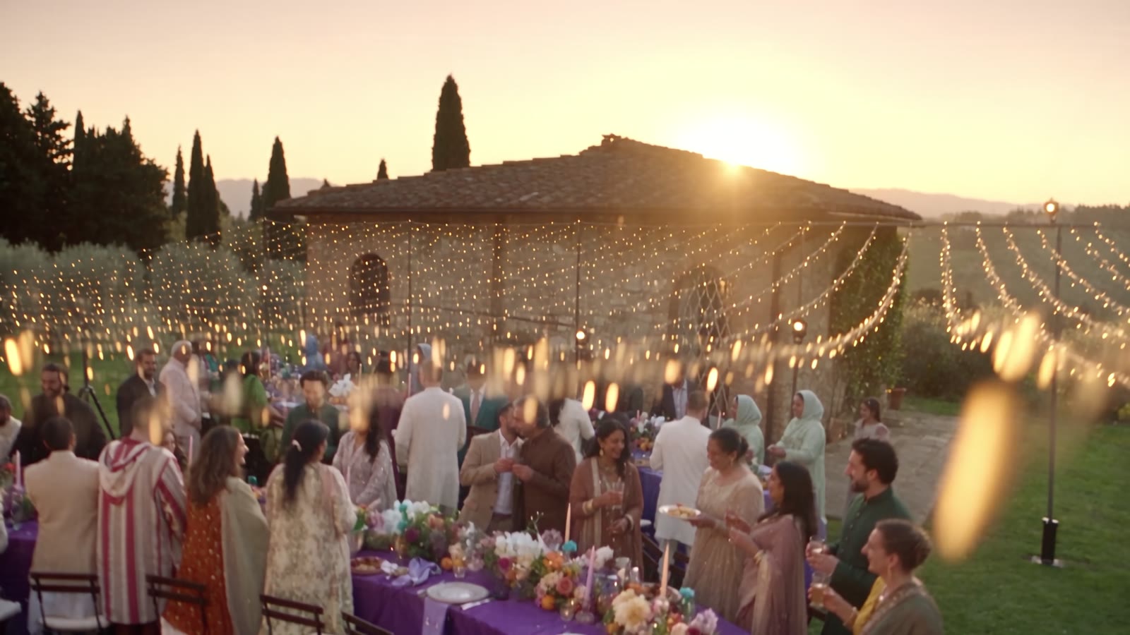 Gli invitati lungo la tavola viola a S sotto i fili di luci al tramonto, la villa alle spalle
