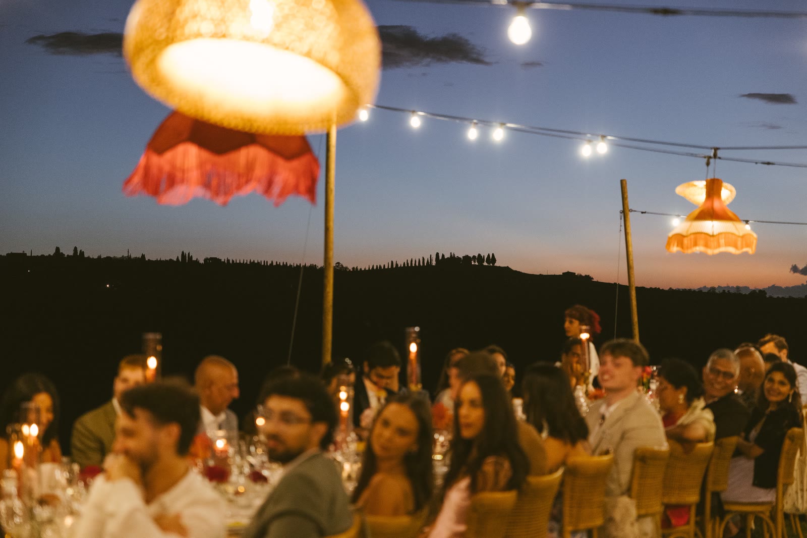 Guests at the long table under the lanterns at night, staff in white serving
